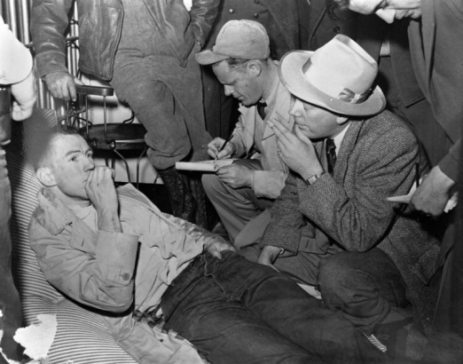 Laying on a wadded, pinstriped prison mattress at the State Penitentiary in Canon City, Colorado, This recaptured escapee has his hand to his mouth. Men write on pads of paper; one wears a baseball cap. A man in a tweed jacket and smoking a cigarette is identified as Red Fenwick; he wears a fedora and square wristwatch. A man in the background leans on a wire chair against a wall.