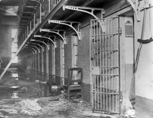 Interior, bracketed metal and pipe railed balconies top cells at the State Penitentiary in Canon City, Colorado. Iron gates are in the brick wall; one in the foreground is open. A writing slate hangs inside the cell, and the facing floor is strewn with blood, debris and a wooden chair. The row-lock is by the door, and a fallen beam leans on the balcony.