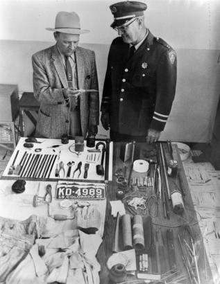 Tools used in a prison break from the State Penitentiary in Canon City, Colorado, include hacksaw blades, drill brace and bits, pliers, a screwdriver, putty knife, batteries, string, wire, and tape. In a Fedora, tweed jacket and tie, associate warden Fred Wyse holds a serrated knife as Captain Chester Yeo, in uniform, looks on. Also on the table are gloves, a spool of fishing line, and Colorado license plate number: "KD- 4989," 1962.