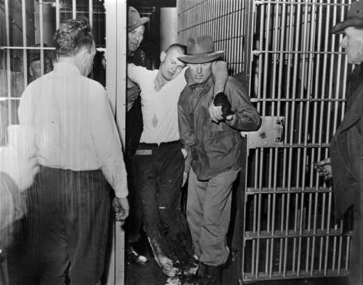 Guards carry a slumping convict (R. L. Freeman, kidnapper) through interior barred gates at the State Penitentiary in Canon City, Colorado. Snow is on their pants, shoes and hats. Men stand in the foreground; one holds a cigar.
