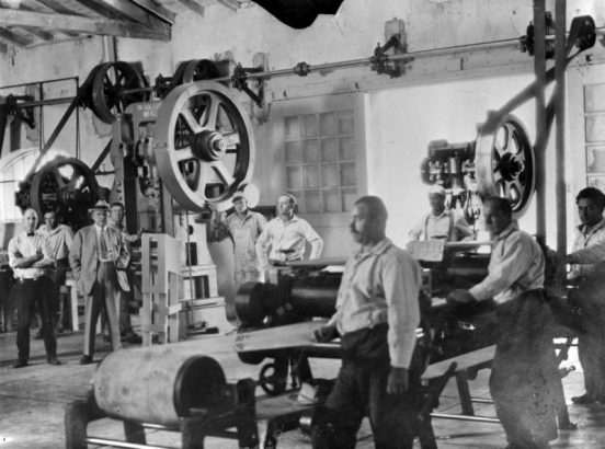 Inmates (one Black) and officials at the State Penitentiary in Canon City, Colorado, pose among metalworking machines with rollers, flywheels and belted pulleys. An axle mounted on an interior wall drives a drill press and a stamp mill, which has "US Toledo No. 57" embossed on its frame.  Track mounted, sliding wooden doors with paned windows are open, and glare comes in from outside. The men are in striped shirts, coveralls, and a suit and tie.