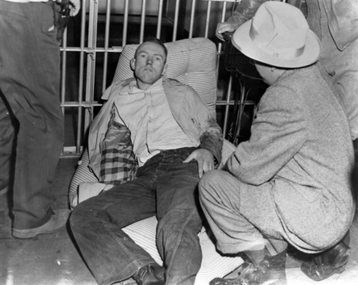 Lying on a wadded, pinstriped mattress, This captured escapee from the State Penitentiary in Canon City, Colorado, appears defiant in his dungarees, shirt and stained jacket. Interior prison bars are behind him; the man squatting in the foreground is identified as Red Fenwick.