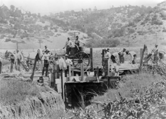Men work at the State Penitentiary in Canon City, Colorado. Plank walls and roof surround an opening fronted by a channel in the foreground. With bags, shovels, and wheelbarrows, inmates are watched by men sitting on barrels in a mule drawn wagon. Power lines, railroad track, and low hills are in the background.