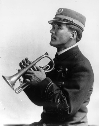 Cornet in hands, the leader of the State Penitentiary Band in Canon City, Colorado poses in profile. His uniform has appliqued braiding, and his cap has a shiny bill and brass band insignia. He wears wire rimmed spectacles.