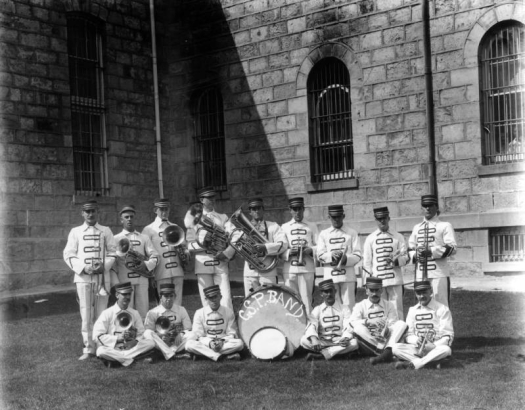 Members of the prisoner's band at the State Penitentiary in Canon City, Colorado, pose with drums, trombones, cornets, and trumpets. Their uniforms are white with rectangles across the fronts, and they wear conductor's hats. Stone Prison buildings with extruded joints and arched, barred windows are behind them.