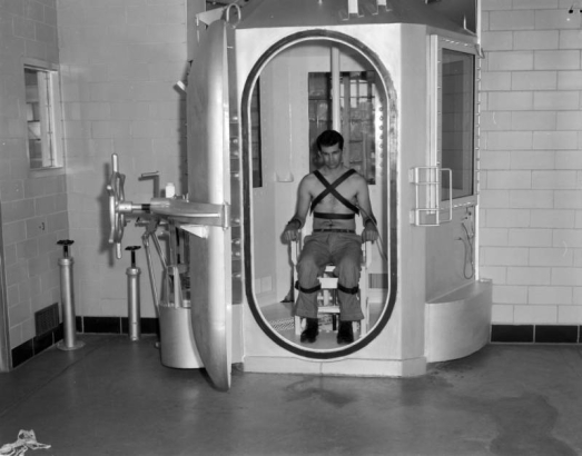 A man poses strapped to a chair in the gas chamber at the State Penitentiary in Canon City, Colorado. He wears pants, belt and shoes; a tube connected to his chest is linked to a stethoscope under a window on the exterior of the unit. The door has a wheel closure; adjacent pipes are topped with wheel valves. A lever and spring are in back, a white plastic jar is atop the door mechanism.