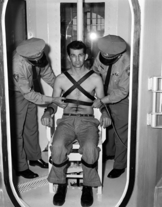 Guards adjust straps on a man posed in the chair in the gas chamber at the State Penitentiary in Canon City, Colorado. He wears pants, belt, shoes, and a tube connected to his chest. Flashbulb glare reflects from interior glass, and a perforated metal conduit shield is on the floor. A hinge and rubber seals are in the foreground.