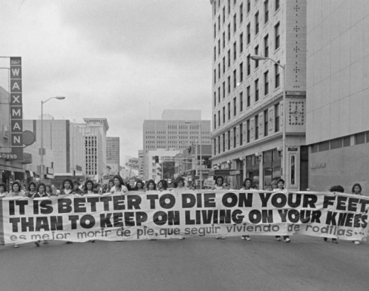 Mexican Americans, members of the Crusade for Justice (La Crusada Para Justicia) march in protest down 15th (Fifteenth) street in Denver, Colorado. They carry a large banner written in both English and Spanish, which reads: "It Is Better to Die On Your Feet Than to Keep On Living On Your Knees," "Es Mejor Morir de Pie, Que Seguir Viviendo de Rodillas."