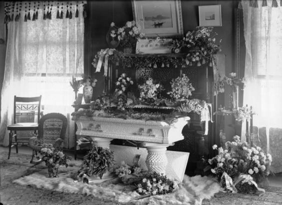 Edwin Felt lies in his coffin in (probably) Denver, Colorado; floral tributes, a piano, a fur rug, and tapestries with tassels surround the child's carved wooden bier.