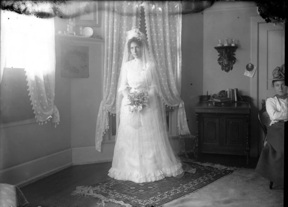 Blanche Roberts poses in possibly a wedding dress with a veil, in (probably) Denver, Colorado. She holds a bouquet of flowers; another woman sits to the side. Decor includes a woven rug and a sconce shelf with a mounted taxidermic antelope and antlers.