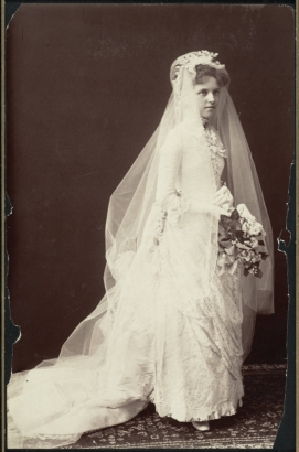 A bride, Mary Virginia King, poses for a studio portrait, probably at Willets Point (Queens County), New York. She wears a long wedding dress with draped lace over a box pleated underskirt, long fitted sleeves, a fitted bodice, gathered or layered lace collar ruffles, gloves, and a long veil crowned with blossoms. A long, silk train is pooled on an oriental rug. A bouquet has leaves and stephanotis blossoms.