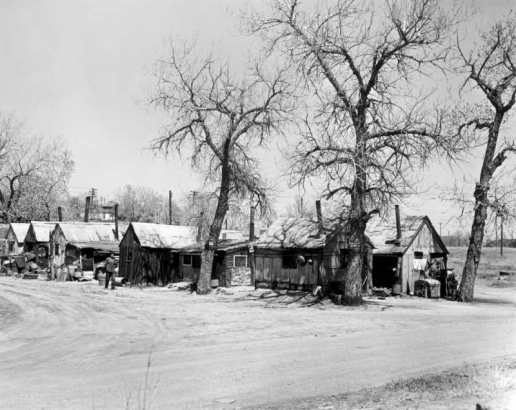 View of shanties on a dirt road near probably 19th (Nineteenth) Avenue and Clay Street in the Jefferson Park neighborhood of Denver, Colorado. Houses are made of wood and corrugated metal and have chimneys and roofs patched with shingles and sheet metal. A man in the road wears a v-neck sweater and a hat and smokes a pipe. Trees, scrap wood, metal pots, and laundry on a clothesline are beside buildings.