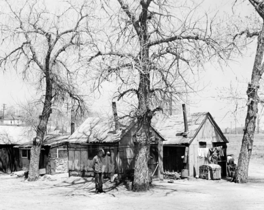 A man wears a v-neck sweater, pants, and a hat and smokes a pipe in front of a shanty at probably 19th (Nineteenth) Avenue between Bryant and Clay Streets in the Jefferson Park neighborhood of Denver, Colorado. Houses are made of wood, corrugated metal, sheet metal, and tar paper. A roof has a scalloped edge at the eves. A pile of bricks, wooden trunks, laundry on a clothesline, and metal buckets are near trees. An address number "2645" is above a door.