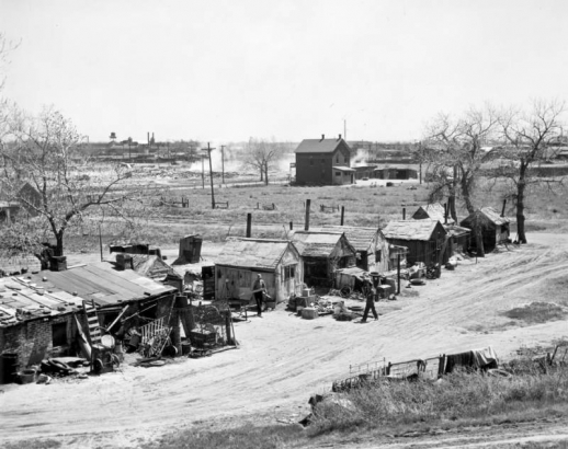 View of a row of shanties at 19th (Nineteenth) Avenue and Clay Street in the Jefferson Park neighborhood of Denver, Colorado. Men stand near buildings made of brick, corrugated metal and tar paper. Shingle roofs are patched with metal or weighted by bricks. Wire fences, tires, bed frames, and wheelbarrows are on the ground. Brick buildings, fenced lots, smokestacks, a water tower, dirt roads, and dust are in the distance.