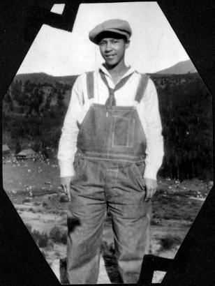 Portrait of Marie L. Anderson (later Marie L. Greenwood), an African American teenager, the young woman poses wearing overalls, tie, shirt and a cap. Camp Nizhoni in Lincoln Hills (Gilpin County), Colorado is in the distance. Shows scattered people on the campgrounds.