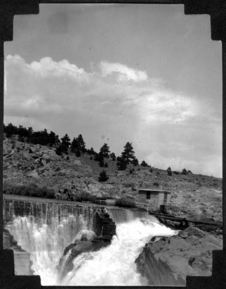 View of the dam at the entrance of Big Thompson Canyon (Larimer County), Colorado. Water flows over the dam, and a small concrete building near large pipes is shown.