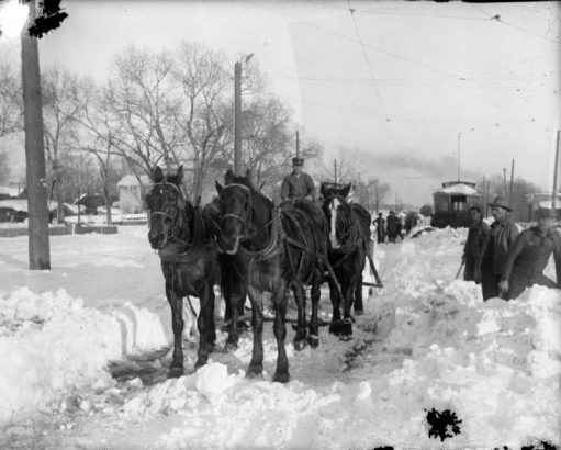 A man rides on a horse-drawn snow plow on Alameda Avenue in the Valverde neighborhood of Denver, Colorado. A group of men with shovels and a trolley car are nearby.