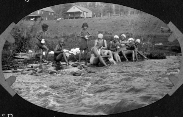 A group of African American campers from Camp Nizhoni bathe on the bank of South Boulder Creek in Lincoln Hills (Gilpin County), Colorado. The teenage girls wash clothes and wear swim suits and caps. Camp buildings are in the distance.