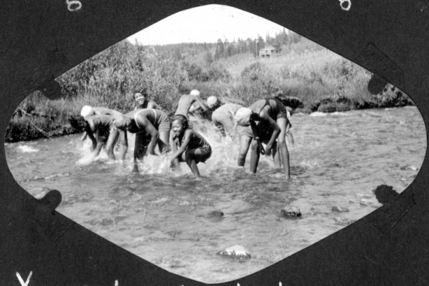 African American teenage campers wear swim suits and swim caps as they splash in South Boulder Creek in Camp Nizhoni in Lincoln Hills (Gilpin County), Colorado. A campground building and flagpole are in the distance.