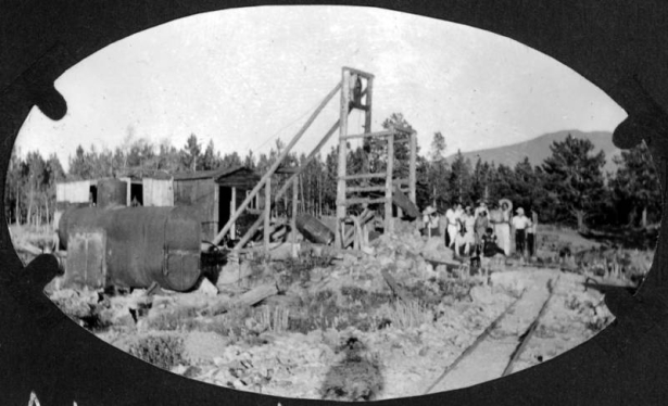 View of the headframe of a tungsten mine and large metal tanks. African American female campers and counselors from Camp Nizhoni stand by the mine, probably in Gilpin County, Colorado. Shows ore car tracks.