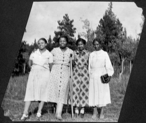 Four African American women, Katherine Patton (Pat), Mary E. Wood (Woody), Marie Anderson ( Andy, later Marie L. Greenwood), and Mrs. Wilson pose together at Camp Nizhoni in Lincoln Hills (Gilpin County), Colorado. Woody has a broken leg and leans on one crutch. They all wear dresses; two of which have a print pattern. Mrs. Wilson holds a purse.