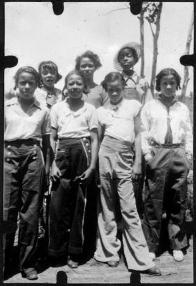 Seven African American teenage campers from Camp Nizhoni pose in Lincoln Hills (Gilpin County), Colorado. They all wear pants and blouses; one has on overalls, one wears a straw hat and three others wear sailor-type pants with large buttons.