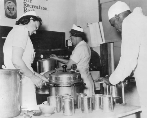 Women and an African American man can food probably for school lunch programs in Denver, Colorado. Shows cans, large pots and pressure cookers.