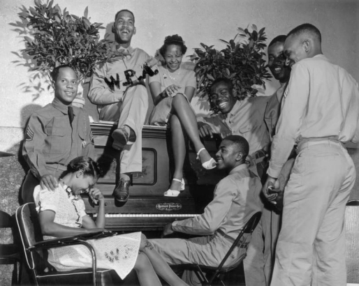 African American soldiers in uniform and women laugh as they stand near, or sit on, a piano at a W.P.A. armed forces center in Denver, Colorado. The women wear dresses, one has on strap heeled shoes.