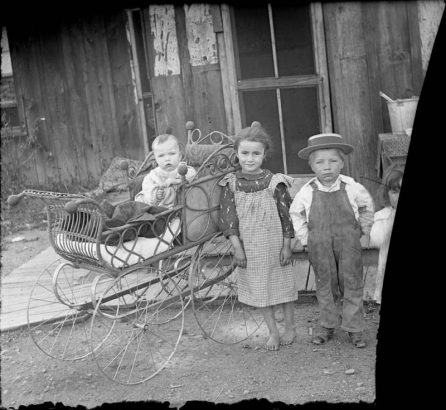 Outdoor portrait of children, they include Lydia Lillybridge (Medin), Montana Lillybridge (Ericson), and a baby in a carriage near a house in Denver, Colorado. The boy wears overalls and a straw hat.