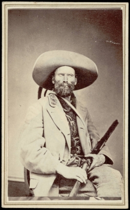 Studio portrait of a man with a beard, rifle, and wide brimmed hat.