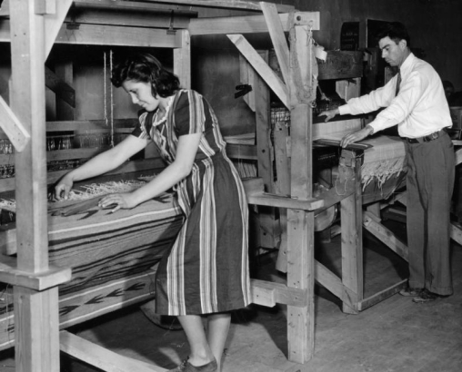 A Hispanic American man and woman work on traditional wooden treadle looms probably in Southern Colorado. The woman works on a Rio Grande style weaving.