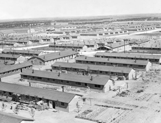 Overview of prefabricated army-style barracks at the Granada Relocation Center, Camp Amache, Prowers County, southeastern Colorado, includes small trees and gardens with rolling prairie in the background.