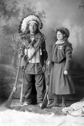 Annie Oakley and unidentified Native American male, both standing, full length studio portrait with painted backdrop; Oakley wears western hat, dress, medals on chest, holding a rifle in left hand; unidentified Native American wears feather headdress, necklace, fur wrapped around shoulders, breastplate, sash, beaded bands on arms, and holds rifle in left hand.