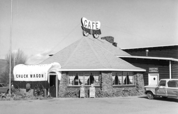 Exterior of the Chuck Wagon Cafe and Cocktail Lounge, Granby, Grand County, Colorado, shows the stone restaurant with pyramidal roof, covered wagon entrance, and painted log back addition. A neon covered wagon  sign tops the roof. Rocky Mountain News and Denver Post newspaper boxes stand at the entrance. Signs in curtained windows read: "Help Wanted," "Come In We're, Open," and "Yes...We Are Open."