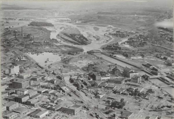 Aerial view of a flooded Arkansas River, damaged buildings and rail yards in Pueblo (Pueblo County), Colorado. Shows the Pueblo Smelter, the Nuckolls Packing Company building, standing water identified as "Death Lake", a flooded Missouri Pacific Railway Company rail yard, Denver and Rio Grande Western and Atchison, Topeka and Santa Fe railroad bridges, and City Hall with a domed cupola.