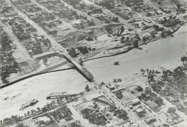 Aerial view of Fountain Creek, a damaged 4th (Fourth) Street bridge, a flooded and destroyed Colorado and Southern Railway bridge in the creek bed, and Denver and Rio Grande Western tracks beside the creek in Pueblo (Pueblo County), Colorado.