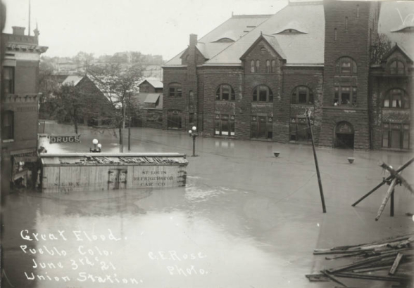 The intersection of B Street and Victoria Avenue is under flood water from the Arkansas River in Pueblo (Pueblo County), Colorado. Shows water in the entrance to the stone Union Depot building, submerged telephone poles and a "St. Louis Refrigerator Car" washed against a drug store building.
