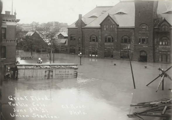 The intersection of B Street and Victoria Avenue is under flood water from the Arkansas River in Pueblo (Pueblo County), Colorado. Shows water in the entrance to the stone Union Depot building, submerged telephone poles and a "St. Louis Refrigerator Car" washed against a drug store building.