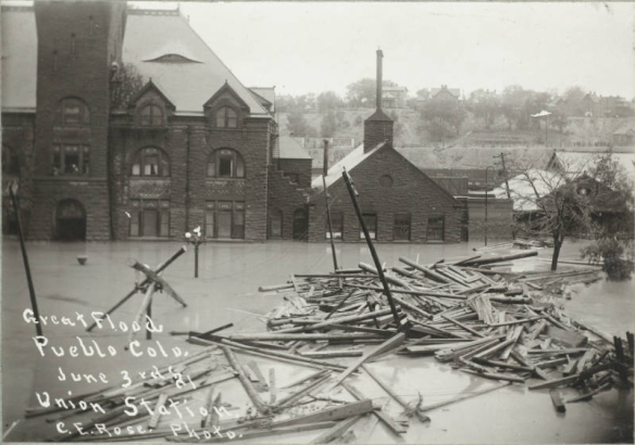View of lumber and debris near the intersection of B Street and Victoria Avenue in Pueblo (Pueblo County), Colorado. Shows water in the entrance to the stone Union Depot building and submerged telephone poles and trees.