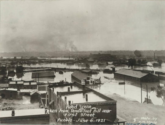 View of the flooded Arkansas River in Pueblo (Pueblo County), Colorado. People stand and look at the destruction from railroad tracks in Goat Hill (Tenderfoot Hill) in northeast Pueblo. Shows a railroad bridge, buildings submerged in water, probably the Nuckolls Packing Company building, and smokestacks in the distance.