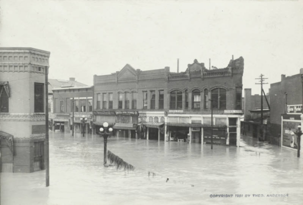 View of flood water and submerged commercial buildings at the intersection of Main Street and Union Avenue in Pueblo (Pueblo County), Colorado. Shows debris on a fallen telephone wire, and signs on businesses that read: "Stark Clothing Co.," "The Wonder Men's Store," "United Shoe Stores Co.," "Central Studio," and "Central Market."