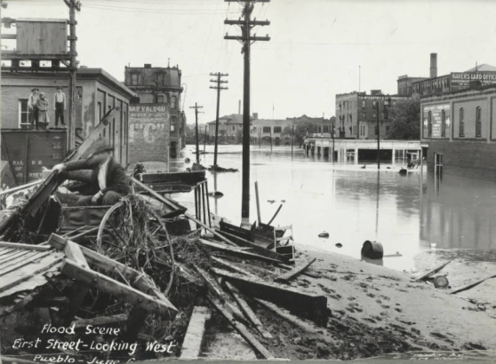 People pose on a freight car and on a pile of debris beside flood water from the Arkansas River on 1st (First) Street in Pueblo (Pueblo County), Colorado. Shows a pile of lumber, a wagon wheel, and flood water on First Street. Brick buildings read: "Haver's Land Office," "T.G. McCarthy and Co. Undertakers," and "Auto Parts & Rubber House, United States Tires, Sales & Service Depot."