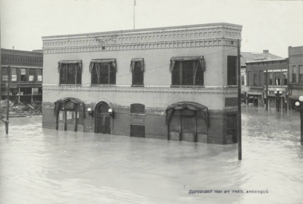 View of a two-story brick building with a flat roof and cloth awnings identified as the "Old White Triangle Block" on Union Avenue and Main Street in Pueblo (Pueblo County), Colorado. Flood water from the Arkansas River surrounds commercial businesses. Shows submerged doorways, windows, street lights, telephone poles, and piles of debris.