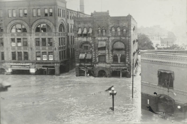 View of Arkansas River flood water at the intersection of Main Street and Union Avenue in Pueblo (Pueblo County), Colorado. Flood water is half way up the first story of stone commercial buildings with arched doorways and windows, and flat roofs. Flood debris is on power lines in the street.