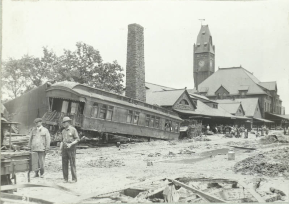 Men stand outside the Union Depot in Pueblo (Pueblo County), Colorado. One man wears a badge. Shows water, mud and debris on the ground, and a flood damaged passenger car wrecked beside the stone depot. Probably a cleaning crew works on mud covered tracks nearby.