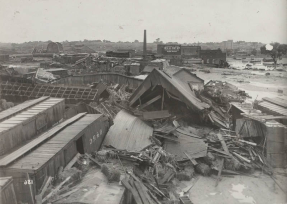 View of wrecked Denver and Rio Grande Western freight cars and frame buildings after the Arkansas River flood in Pueblo (Pueblo County), Colorado. Piles of lumber and debris, mud and silt are in the Walker rail yard. A smokestack is beside a building that reads: "This Property for Sale or Rent, apply to H.C. Borndruck, Phone 120."