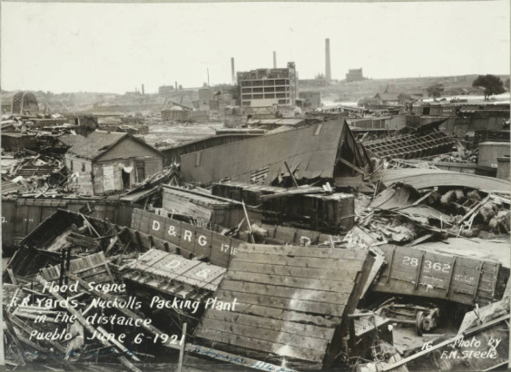 Wrecked buildings and overturned Denver and Rio Grande Western freight cars are in the Walker rail yard after the Arkansas River flood in Pueblo (Pueblo County), Colorado. Shows smokestacks and the Nuckolls Packing Company buildings in the distance.