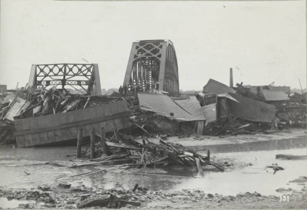 View of an Atchison, Topeka & Santa Fe railroad bridge and a wrecked Denver & Rio Grande Western railroad bridge with piles of wreckage from the Arkansas River flood in Pueblo (Pueblo County), Colorado. Men stand and pose on piles of debris and rubble from flooded buildings and rail yards.