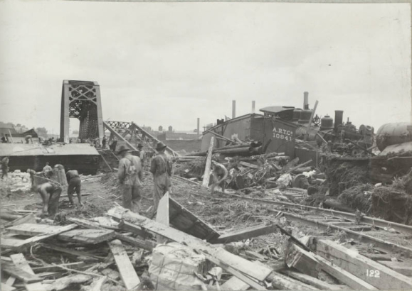 Men work to clear rubble piled along the flooded Arkansas River in Pueblo (Pueblo County), Colorado. Freight cars, a locomotive, hay bales, tracks and lumber are scattered near Atchison Topeka & Santa Fe and Denver & Rio Grande Western railroad bridges.