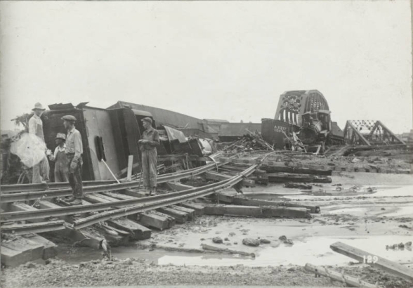 Men in overalls or work clothes and hats stand on washed out railroad tracks beside piles of rubble from the Arkansas River flood in Pueblo (Pueblo County), Colorado. Atchison Topeka & Santa Fe and Denver & Rio Grande Western railroad bridges are in the distance. Shows wrecked and overturned locomotives and railroad cars.